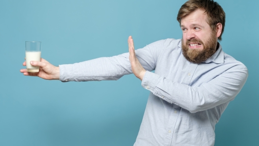 Lactose intolerance. Dairy intolerant man making a stop gesture glass of milk, which is holding in hand. Isolated on a blue background.