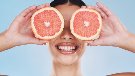 Beautiful young mixed race woman with a grapefruit isolated in studio against a blue background. Her skincare regime keeps her fresh. For glowing skin, eat healthy. Packed with vitamins and nutrients