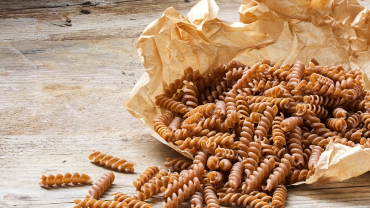 wholemeal pasta fusilli from organic whole grain spelt falling from a paper bag on a rustic wooden table, copy space