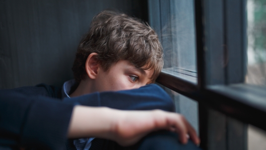 Pensive sad boy teenager with blue eyes in a blue shirt and jeans sitting at the window and closes his face with his hands.