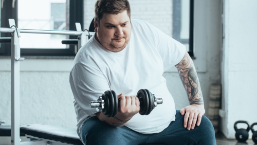 Overweight tattooed man sitting and training with dumbbell at sports center