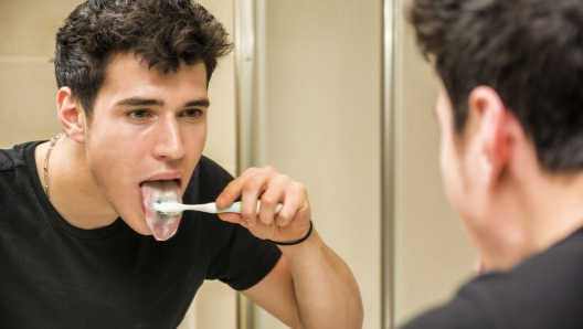 Headshot of attractive young man brushing teeth and cleaning tongue with toothbrush, looking at himself in mirror