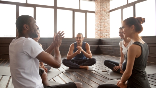 Excited african american man talking to sporty happy diverse friends sitting on mat in studio, happy black yoga instructor or life coach motivating young people having fun at seminar training class