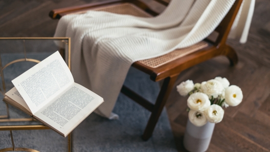 Concept of living room with hygge design. White knitted soft blanket on wood chair, book on glass side table and blooming flowers in vase on the floor