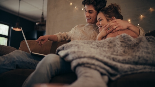 Young couple sitting on sofa at home in winter and using laptop computer. Cozy couple in hygge house with laptop.