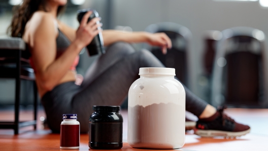 Selective focus on protein powder and multivitamins jars on the gym floor with a sporty woman in background.