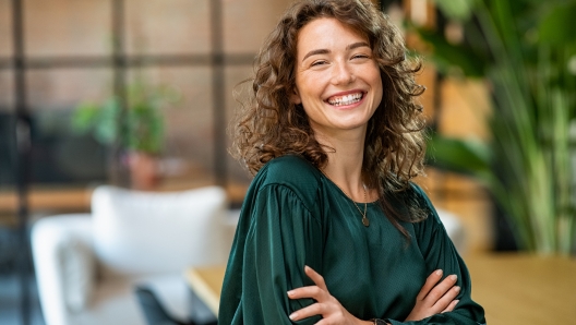 Portrait of young smiling woman looking at camera with crossed arms. Happy girl standing in creative office. Successful businesswoman standing in office with copy space.