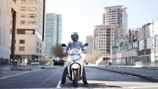 Wide shot of a young motorcyclist stopped at a traffic light in Barcelona. The man riding his scooter through the city on a large avenue lined with skyscrapers is waiting at the traffic light.