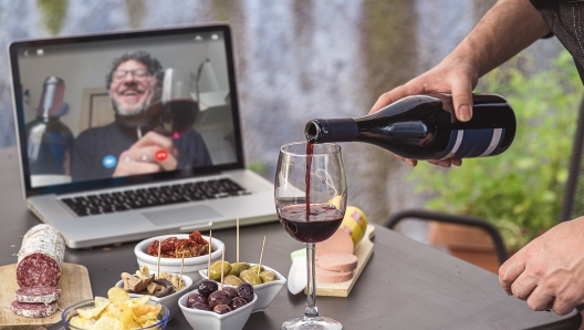 Lockdown aperitif video call party. Adult men are making a pre-meal aperitif with snacks, wine, and Italian appetizers together at home using teleconference platform apps during COVID-19 restrictions