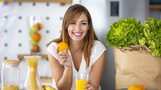 Young joyful woman drinking orange juice and standing near a kitchen table. Close up of a woman drinking juice in her kitchen. Fit smiling young woman preparing healthy fruit juice