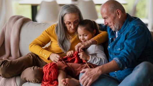 A little girl sitting on sofa with her grandparents and learning to knit indoors at home.