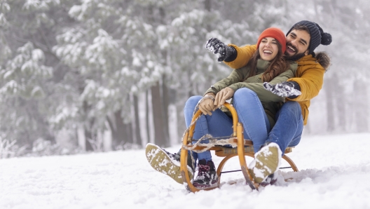 Beautiful young couple in love having fun spending winter vacation in mountains, sitting and hugging on sled, sliding down the hill in the snow