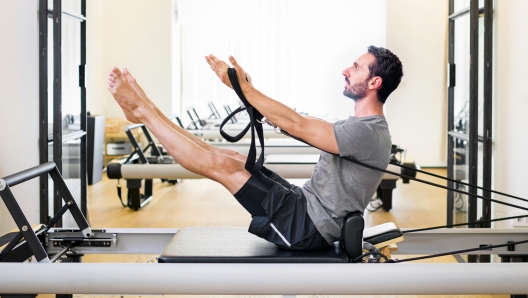 Fit muscular man doing a teaser pilates exercise on a reformer in a gym to strengthen his core and abdominal muscles in a side view with copyspace