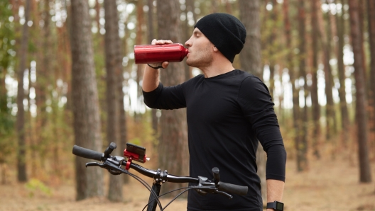 Image of young thirsty athletic sportsman drinking water with closed eyes, having peaceful facial expression, standing in profile, holding his bicycle, being in forest during training. Sport concept.