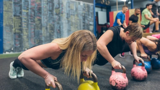 Sportswomen doing push-ups with kettlebells with mates doing box jumps in background