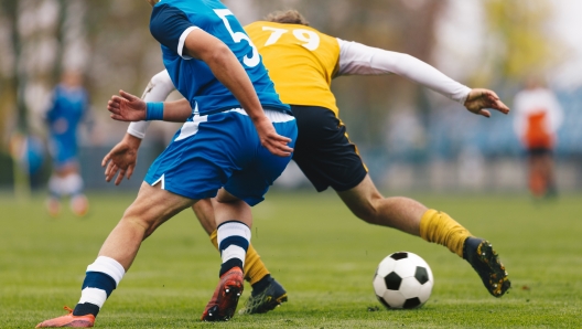 Football Players Compete in Tournament Match in a Duel. Football Game on Summer Sunny Day. Adul Soccer Players in Blue and Yellow Jersey Uniforms Running Classic Soccer Ball on Grass Pitch