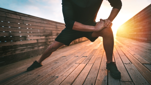 Young slim sporty man stretching leg muscles and warming up for training outdoors at sunset or sunrise. Athletic man in black sportswear doing fitness stretching exercises. High quality photo