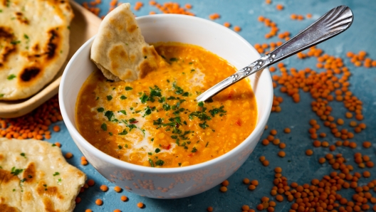 Red lentil soup with coconut milk and curry accompanied by naan bread sorrounded by uncooked lentils on a blue background