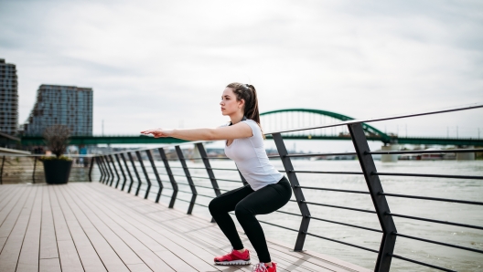 Side portrait of young woman doing squat exercises on a river bank.