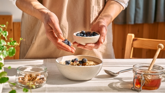 Woman preparing healthy dieting vegan nutritious breakfast. Female hand putting blueberries in the bowl with oatmeal porridge with walnuts and honey.