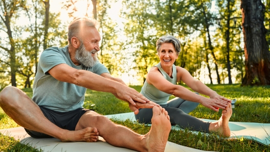 Flexible exercises for body. Sporty man and woman with grey hair stretching on yoga mats with hands to one leg during outdoors workout. Happy married couple with bare feet warming up together at park.