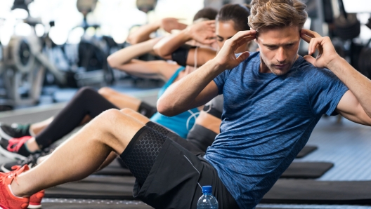 Muscular guy doing sit ups at gym with other people in background. Young athlete doing stomach workout in modern gym. Handsome fit man doing crunches at gym.