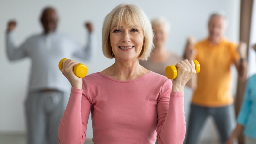 Selective focus on cheerful senior lady posing with fitness tools, multiracial group of healthy elderly people in sportswear doing strength building workout with dumbbells at gym