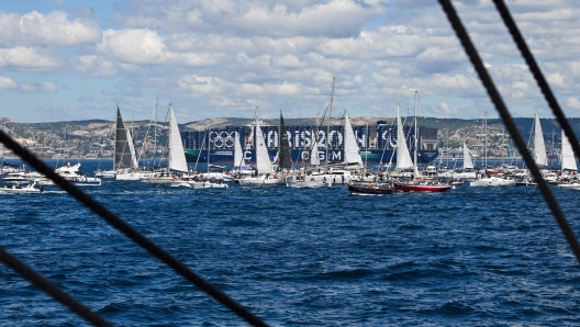 This photograph taken from the French 19th-century three-masted barque Belem shows containers with the logo of Paris 2024 Olympic Games, on the Greenland container ship of the CMA CGM and the boats parade as the Belem sails in the bay of Marseille, in the Mediterranean Sea, on May 8, 2024, before landing with the Olympic torch, ahead of the Paris 2024 Olympic and Paralympic Games. The Belem is set to reach Marseille on May 8 and ten thousand torchbearers will then carry the flame across 64 French territories. It will travel through more than 450 towns and cities, and dozens of tourist attractions during its 12,000-kilometre (7,500-mile) journey through mainland France and overseas French territories in the Caribbean, Indian Ocean and Pacific. On July 26 it will form the centrepiece of the Paris Olympics opening ceremony. (Photo by NICOLAS TUCAT / AFP)