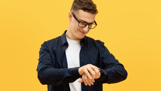 Time management concept. Positive young guy in eyeglasses checking time, looking at smart watch on his wrist and smiling, isolated on yellow studio background