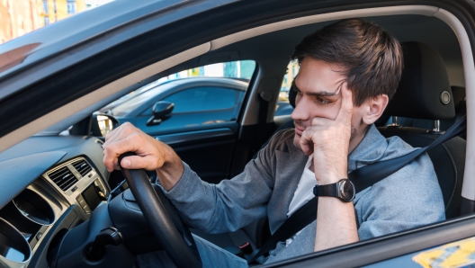 Annoyed tired young man is riding driving car. Businessman is late for meeting. Driver brunette in grey suit stuck in traffic jam. Stressful situations on roads and fast rhythm in modern city.