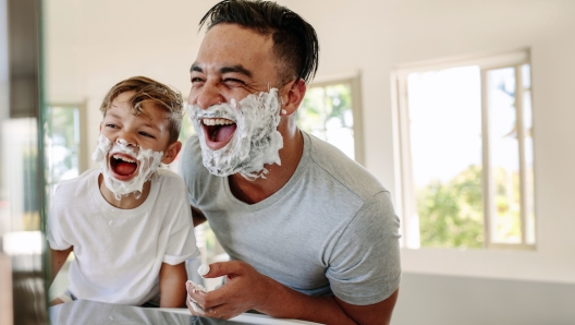 Father and son having fun in a bathroom, laughing happily with shaving foam on their faces. Young single dad taking a moment to bond and share moments of joy with his boy on father's day.