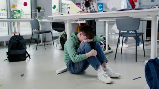 Depressed lonely boy sitting under desk in classroom with classmates playing on background having break