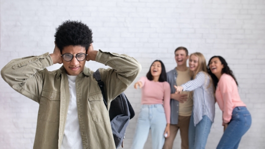 Bullying victim, depressed student suffers from school humiliation. Sad young african american guy in glasses closes ears, as not to hear laughter of international people, studio shot, empty space