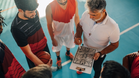 High angle view of a mature confident male coach discussing game tactics with his multiracial basketball team using a coach clipboard and explaining game plan and drawing defense. Copy space.