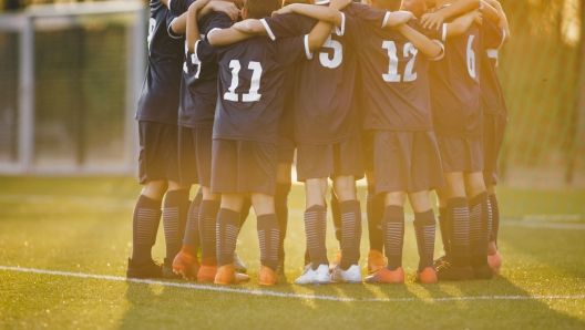 Group of children huddling with coach. Summer sunset at the stadium in the background. Youth soccer football team group photo. Happy boys soccer players kicking tournament. School boys in blue jerseys