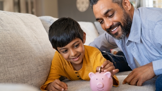 Happy indian son saving money in piggy bank with father. Lovely ethnic father teaching to little boy importance of saving money for future. Smiling middle eastern kid adding coin in piggybank while lying on couch with dad at home.