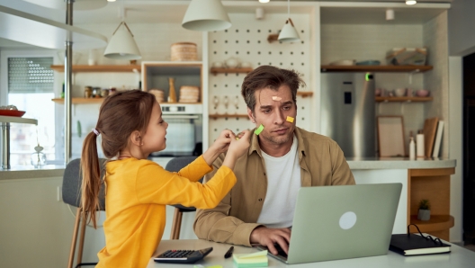 Tired man working on a laptop with his face being covered in sticky notes by his daughter.