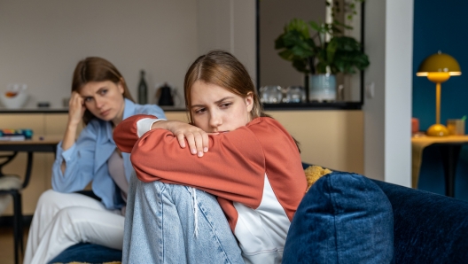 Frustrated teenage girl sits hugging his knees, looks away. Mom sits next daughter and comforts after quarrel. Communication problems between parent and kids.Ignoring children problems.Bulling school