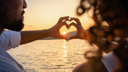 View from back of a young beautiful couple doing heart shape with fingers looking at the sun setting behind the mountains, reflecting light in the sea water. Valentine day love concept. Focus on hands