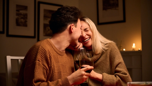 Happy young couple in love hugging, laughing, drinking wine, enjoying talking, having fun together celebrating Valentines day dining at home, having romantic dinner date with candles sitting at table.