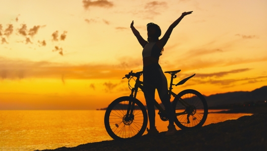 Silhouette of active one free happy joyful carefree inspired woman bicycler with open arms standing alone with bicycle outdoors against sunset sky