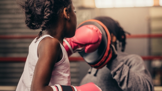 Girl wearing boxing gloves straining with her coach. Boxing kid practicing punches on a punching pad with her coach.