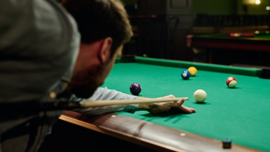 Focus on hand of young man holding cue on billiard table with green top while bending over it and concentrating on one of balls