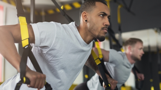 Young men performing TRX training in gym. Muscular guys doing exercise with elastic rope, copy space, closeup