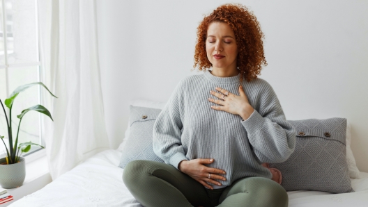 Portrait of plus-size curly redhead female in casual clothes doing pranayama technique sitting on bed next to window with closed eyes, putting hands on chest, breathing slowly and deeply