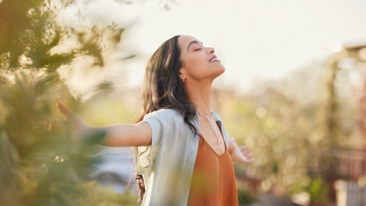 Young latin woman with arms outstretched breathing in fresh air during sunrise at the balcony. Healthy girl enjoying nature while meditating during morning with open arms and closed eyes. Mindful woman enjoying morning ritual while relaxing in outdoor park.