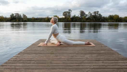 Mature woman practicing yoga, doing exercises on four limbs, push-ups from the floor, Half pose of the royal pigeon, Eka Pada Rajakapotasana, training, dressed in sports clothes. outdoor nature background. Autumn evening by the river.