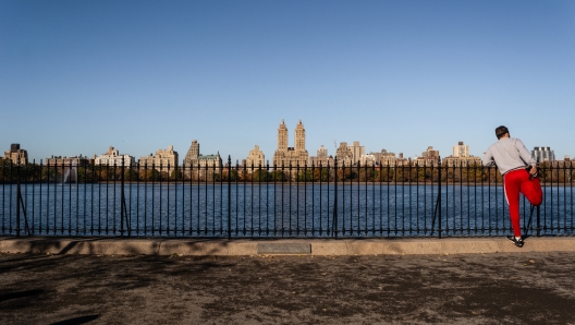 A runner stretching along the path of running path of the Central Park Reservoir on a fall day with the Upper West Side skyline in the background.