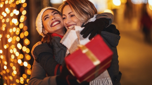 Cheerful two young women are having fun in the city street at the Christmas night. They are laughing, hugging and giving presents to each other.