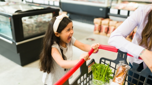 Angry little kid screaming and throwing a tantrum while grocery shopping with her mom at the supermarket because she won't buy her candy